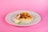 A taco topped with pieces of meat, diced mango, chopped vegetables, and herbs sits on a white plate. A lime wedge is placed beside the taco. The background is a solid pink color.