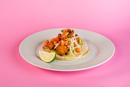 A taco is placed on a white plate against a pink background. The taco is filled with fried items that look like fish, topped with diced tomatoes, shredded lettuce, and white cheese. A lime wedge accompanies the taco to the side.