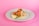 A taco is placed on a white plate against a pink background. The taco is filled with fried items that look like fish, topped with diced tomatoes, shredded lettuce, and white cheese. A lime wedge accompanies the taco to the side.