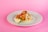 A taco is placed on a white plate against a pink background. The taco is filled with fried items that look like fish, topped with diced tomatoes, shredded lettuce, and white cheese. A lime wedge accompanies the taco to the side.