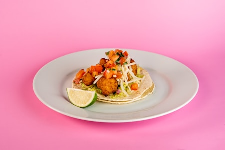 A taco is placed on a white plate against a pink background. The taco is filled with fried items that look like fish, topped with diced tomatoes, shredded lettuce, and white cheese. A lime wedge accompanies the taco to the side.