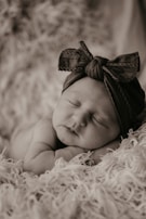 A smiling baby wearing a pastel bow headband sitting on a cozy blanket