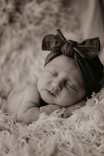 A sleeping baby with a headband featuring a large bow lies on a fluffy blanket. The scene is peaceful and serene, captured in a soft monochrome tone that enhances the texture of the blanket and the gentle expression on the baby's face.