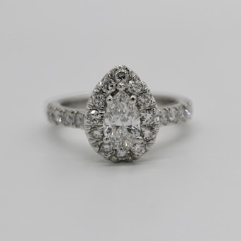 A pear-shaped diamond engagement ring features a central pear-cut diamond surrounded by smaller round diamonds. The band is also adorned with a line of sparkling diamonds, providing an elegant and luxurious appearance.
