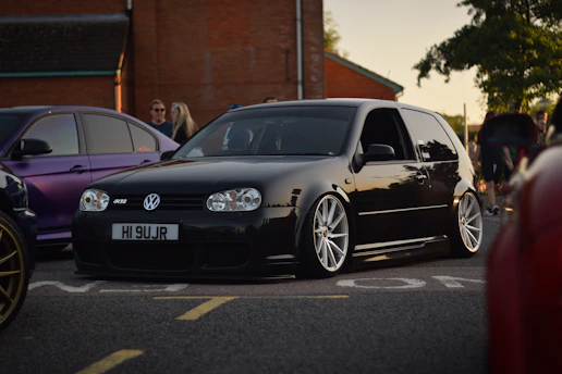 A sleek black car with shiny silver alloy wheels is parked in a parking lot. The vehicle is a Volkswagen, recognized by its distinct logo on the front grille. The scene is set during the day, with several other cars and people in the background, suggesting a gathering or car meet. The surroundings include a brick building and green trees under clear skies.