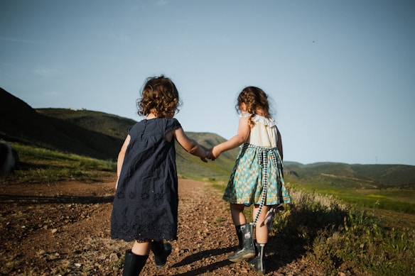 Two young children are walking hand in hand along a dirt path surrounded by green hills, with mountains in the background. The children are dressed in summer clothing and wearing boots, suggesting an outdoor adventure. The sky is clear and blue, indicating sunny weather.