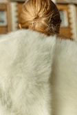 A person with blonde hair is seen from behind, wearing a luxurious white fur garment. The background is softly blurred, likely depicting a building with arched windows.