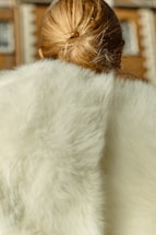 A person with blonde hair is seen from behind, wearing a luxurious white fur garment. The background is softly blurred, likely depicting a building with arched windows.