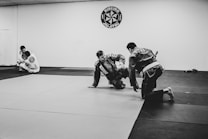 Two people are practicing Brazilian Jiu-Jitsu on a mat in a dojo, while another person observes from the side. They are wearing traditional martial arts uniforms called gi. The environment suggests a training session with a focus on grappling techniques.