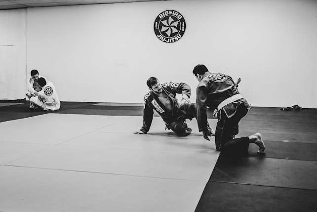 Two people are practicing Brazilian Jiu-Jitsu on a mat in a dojo, while another person observes from the side. They are wearing traditional martial arts uniforms called gi. The environment suggests a training session with a focus on grappling techniques.