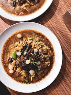 A plate of rich, dark brown stew with tender pieces of meat, grains, and small white onions is garnished with fresh green herbs. The dish is served on a white plate and placed on a wooden table, showing a warm, inviting presentation.