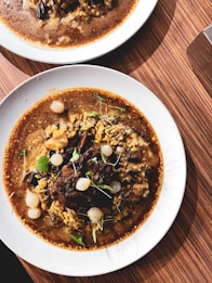 A plate of rich, dark brown stew with tender pieces of meat, grains, and small white onions is garnished with fresh green herbs. The dish is served on a white plate and placed on a wooden table, showing a warm, inviting presentation.