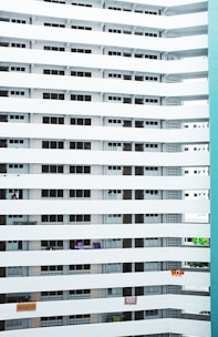 A modern high-rise residential building with multiple floors and balconies. The architectural style is characterized by horizontal white beams separating the floors, with uniform windows and balcony spaces. Some balconies have clothes or towels hanging, suggesting everyday life.