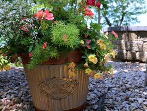 A metal planter labeled 'Flowers & Garden' is filled with an assortment of vibrant flowers and green foliage. The flowers are a mix of pink, red, yellow, and white blossoms, surrounded by lush greenery. The planter is placed on a bed of small, gray pebbles with a stone wall visible in the background.
