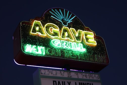 A neon sign glowing against a dark sky, featuring the words 'AGAVE GRILL' prominently in bright yellow and green. Below 'AGAVE,' 'GRILL' is displayed in green, with additional text referring to a restaurant that is less discernible but still illuminated. An agave plant graphic is depicted above the text.