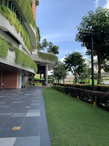 A modern building features vertical gardens with lush green plants growing on the exterior. The pathway beside the building is lined with well-maintained grass, bushes, and trees, creating an urban garden atmosphere. The sky is clear and blue, adding to the serene ambiance. Several square tiles form the pathway with a few decorative elements, enhancing the contemporary architectural design.