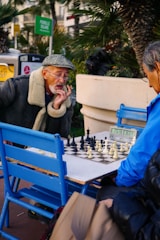 Two elderly men are engaged in an intense game of chess at an outdoor setting. One man is wearing a green flat cap and a coat with a fur collar, appearing deep in thought as he studies the board. The other man is dressed in a bright blue jacket, concentrating on the game. The scene is surrounded by greenery and some urban elements like a sign and a trash bin in the background.
