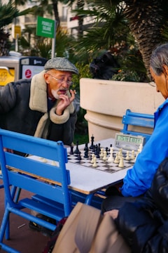 Two elderly men are engaged in an intense game of chess at an outdoor setting. One man is wearing a green flat cap and a coat with a fur collar, appearing deep in thought as he studies the board. The other man is dressed in a bright blue jacket, concentrating on the game. The scene is surrounded by greenery and some urban elements like a sign and a trash bin in the background.