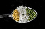 A close-up view of a spoon containing an assortment of grains and legumes. The spoon holds three distinct sections: green mung beans, white basmati rice, and yellow split peas, centered by a single black peppercorn. The background is a dark surface, which highlights the textures and colors of the ingredients.