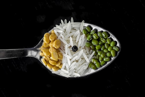 A close-up view of a spoon containing an assortment of grains and legumes. The spoon holds three distinct sections: green mung beans, white basmati rice, and yellow split peas, centered by a single black peppercorn. The background is a dark surface, which highlights the textures and colors of the ingredients.