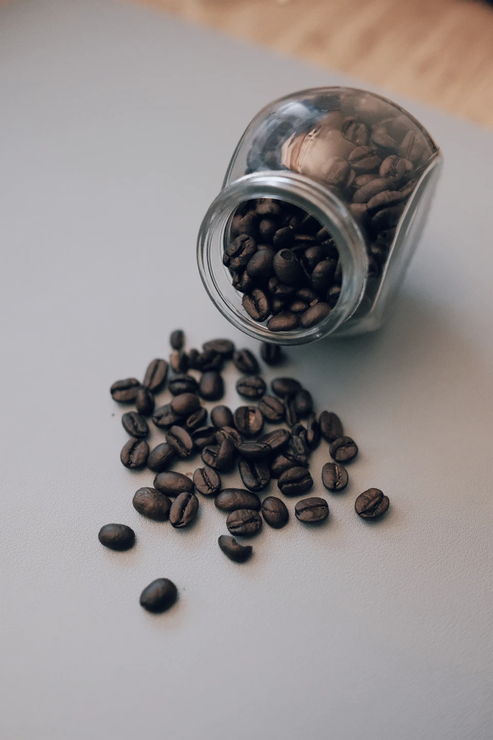 The image shows a glass jar filled with coffee beans, placed on a gray surface. The jar is slightly tilted to the left and has a small amount of coffee spilled out onto the table below it. The coffee beans are scattered around the jar in various sizes and positions, creating an interesting pattern against the transparent glass.