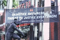 A large banner displaying a quote by Martin Luther King Jr. is hanging between two tall palm trees in front of a brick building. The quote on the banner reads, 'Injustice anywhere is a threat to justice everywhere.' At the base, there is a statue of a man in a uniform, presumably a baseball player, posed as if running or sliding base.