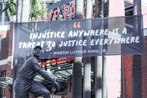 A large banner displaying a quote by Martin Luther King Jr. is hanging between two tall palm trees in front of a brick building. The quote on the banner reads, 'Injustice anywhere is a threat to justice everywhere.' At the base, there is a statue of a man in a uniform, presumably a baseball player, posed as if running or sliding base.