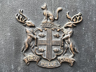 A heraldic emblem featuring a cat at the top, two deer on either side, and a shield in the center. The shield is divided into four sections, with snail-like creatures in the top and bottom left sections and two vertical stripes in the right sections. Below the shield, a banner displays the words 'PRO PELLE CUTEM.' The design is a metallic or bronze color on a textured background.
