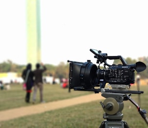 A professional video camera mounted on a tripod sits in the foreground, positioned outdoors in a grassy area. In the blurred background, two people are standing together, seemingly talking or interacting. The overall scene suggests a filming setup in a park or open space.
