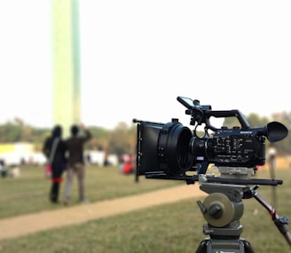 A professional video camera mounted on a tripod sits in the foreground, positioned outdoors in a grassy area. In the blurred background, two people are standing together, seemingly talking or interacting. The overall scene suggests a filming setup in a park or open space.