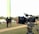 A professional video camera mounted on a tripod sits in the foreground, positioned outdoors in a grassy area. In the blurred background, two people are standing together, seemingly talking or interacting. The overall scene suggests a filming setup in a park or open space.