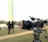 A professional video camera mounted on a tripod sits in the foreground, positioned outdoors in a grassy area. In the blurred background, two people are standing together, seemingly talking or interacting. The overall scene suggests a filming setup in a park or open space.