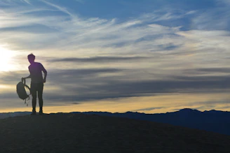 A sunset silhouette of a traveler standing on a mountain peak, gazing at the horizon with a laptop bag slung over their shoulder.