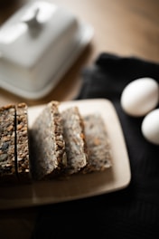 Slices of a whole grain bread loaf are placed on a light-colored cutting board. The bread appears dense and is filled with various seeds and grains. Nearby, two white eggs rest on a dark cloth, while a butter dish is slightly blurred in the background, creating a soft, warm atmosphere.