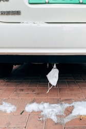 A disposable face mask is hanging from the rear of a vehicle parked on a red brick pavement. Melted patches of snow are scattered across the ground along with some cigarette butts.