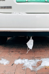 A disposable face mask is hanging from the rear of a vehicle parked on a red brick pavement. Melted patches of snow are scattered across the ground along with some cigarette butts.
