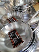 Stacks of stainless steel frying pans with labels are neatly displayed, highlighting their shiny surfaces and sturdy handles.