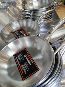 Stacks of stainless steel frying pans with labels are neatly displayed, highlighting their shiny surfaces and sturdy handles.