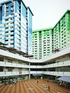 Two tall residential buildings with blue and green exteriors border an open courtyard with a checkered floor pattern. Small shops and food stalls line the ground floor, and several people walk through the area.