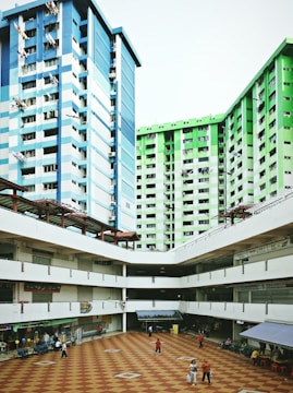Two tall residential buildings with blue and green exteriors border an open courtyard with a checkered floor pattern. Small shops and food stalls line the ground floor, and several people walk through the area.