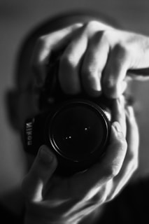 A black-and-white photograph of hands holding a camera, capturing a close-up view of the lens with fingers wrapped around it. The focus is on the hands and the camera lens, while the background is blurred.