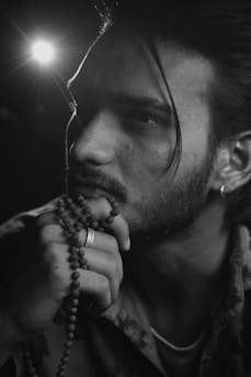 A contemplative man with long hair and facial hair holds prayer beads, with a pensive expression on his face. He wears a ring on one of his fingers and earrings. The lighting in the image is dramatic, casting shadows and highlighting his features.