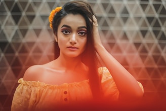 A young woman with long dark hair adorned with a yellow flower stands against a geometric tiled background. She is wearing an off-the-shoulder orange top and gazes directly at the camera with a calm expression. A soft red glow covers the bottom of the image, adding warmth.