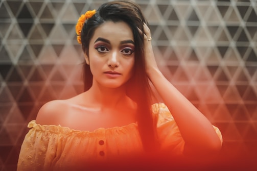 A young woman with long dark hair adorned with a yellow flower stands against a geometric tiled background. She is wearing an off-the-shoulder orange top and gazes directly at the camera with a calm expression. A soft red glow covers the bottom of the image, adding warmth.
