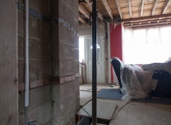A partially constructed or renovated interior space featuring exposed brick walls and wooden ceilings. A white door is visible in the background next to a red painted wall. A plastic sheet covers furniture on the right side, and there are construction materials scattered across the floor.