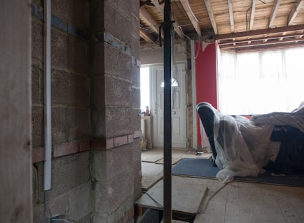 A partially constructed or renovated interior space featuring exposed brick walls and wooden ceilings. A white door is visible in the background next to a red painted wall. A plastic sheet covers furniture on the right side, and there are construction materials scattered across the floor.
