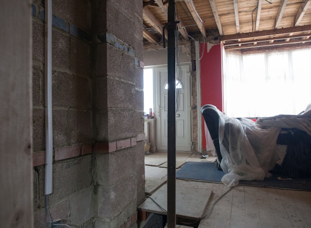 A partially constructed or renovated interior space featuring exposed brick walls and wooden ceilings. A white door is visible in the background next to a red painted wall. A plastic sheet covers furniture on the right side, and there are construction materials scattered across the floor.