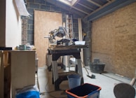 A workshop or construction area has a saw mounted on a table surrounded by various tools and materials. The room has unfinished wooden paneling and a partially constructed ceiling, with a ladder leaning against the wall. Items like buckets and a broom are scattered around, indicating ongoing work.