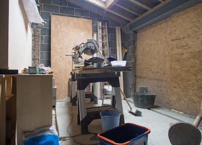 A workshop or construction area has a saw mounted on a table surrounded by various tools and materials. The room has unfinished wooden paneling and a partially constructed ceiling, with a ladder leaning against the wall. Items like buckets and a broom are scattered around, indicating ongoing work.