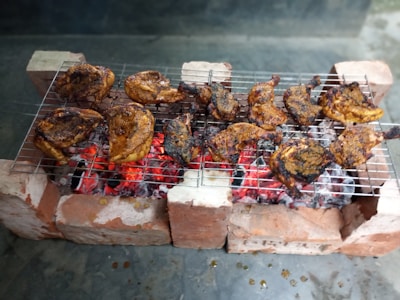 Grilled chicken pieces are arranged on a metal grate over glowing red charcoal, supported by bricks on either side. The chicken is seasoned with spices, giving it a charred, golden brown appearance.